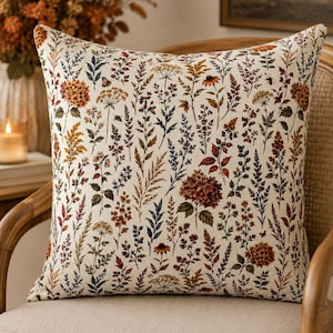 May include: A square throw cushion featuring an off-white background with a dense, repeating botanical print. The pattern includes various wildflowers, grasses, berries, and hydrangea blossoms in shades of rust, orange, brown, and muted blue-green.