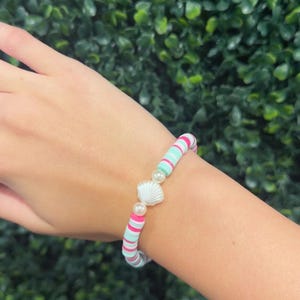 May include: A stretch bracelet featuring flat circular beads in alternating shades of hot pink, light mint green, and white. The bracelet is centered with a white seashell charm flanked by two small, round, cream-colored faux pearl beads.