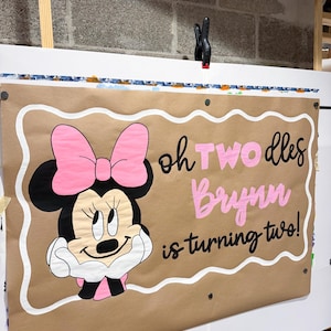 Pode incluir: Uma faixa retangular de papel pardo com um rato de desenho animado com um la&ccedil;o rosa e luvas brancas. O texto diz "oh TWO dles Brynn is turning two!" em letras pretas e rosas. A faixa tem uma borda branca recortada e ilh&oacute;s de metal.