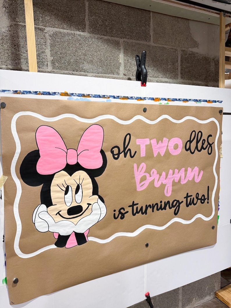 Pode incluir: Uma faixa retangular de papel pardo com um rato de desenho animado com um la&ccedil;o rosa e luvas brancas. O texto diz "oh TWO dles Brynn is turning two!" em letras pretas e rosas. A faixa tem uma borda branca recortada e ilh&oacute;s de metal.