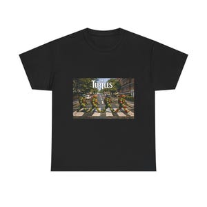 Könnte beinhalten: Ein schwarzes Kurzarm-T-Shirt mit einem Grafikdruck von vier anthropomorphen Schildkröten, die über einen Zebrastreifen auf einer Straße gehen. Über den Schildkröten steht der Text THE TURTLES in weißen Großbuchstaben vor Bäumen.
