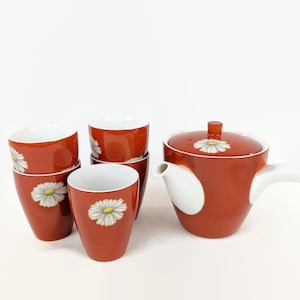 May include: A set of five red and white teacups and a teapot with a daisy design. The teapot has a white handle and a white spout. The teacups are stacked in pairs.