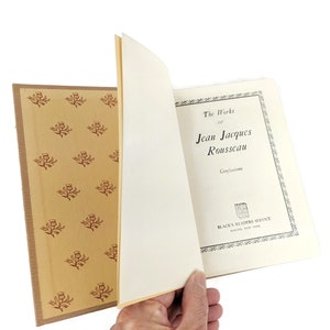 Pode incluir: Um livro vintage intitulado "The Works of Jean Jacques Rousseau" com o subt&iacute;tulo "Confessions". A capa do livro &eacute; branca com texto preto e uma borda preta. O livro est&aacute; aberto para revelar uma p&aacute;gina amarelada com um padr&atilde;o decorativo de flores vermelhas sobre um fundo bege.
