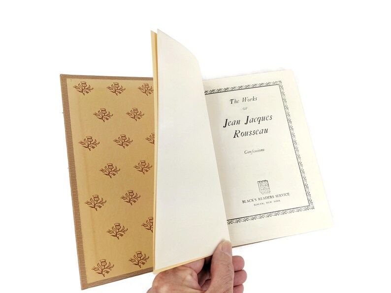 Pode incluir: Um livro vintage intitulado "The Works of Jean Jacques Rousseau" com o subt&iacute;tulo "Confessions". A capa do livro &eacute; branca com texto preto e uma borda preta. O livro est&aacute; aberto para revelar uma p&aacute;gina amarelada com um padr&atilde;o decorativo de flores vermelhas sobre um fundo bege.