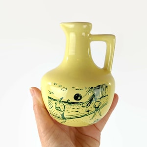 May include: A yellow ceramic pitcher with a green design of people on a beach. The pitcher has a handle and a rounded body.