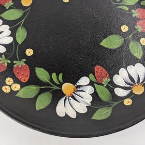 May include: Black plate with a hand-painted floral design. White daisies, red strawberries, and yellow flowers are arranged in a circular pattern around the edge of the plate. Green leaves and stems complete the design.