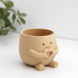 May include: A light tan, matte-finish planter shaped like a rounded character with a smiling face. The character has two arms positioned in front of its chest to form a heart shape with its hands and two small, stubby legs extending forward.