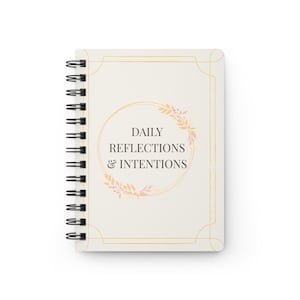 Puede incluir: Un cuaderno de espiral con una cubierta de color crema que presenta un borde dorado fino. El centro muestra el texto DAILY REFLECTIONS & INTENTIONS en negro, rodeado por un marco circular dorado decorado con delicadas hojas color melocotón.