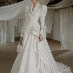 Peut inclure: Une robe longue blanc cassé avec un motif jacquard texturé, dotée de manches longues à épaules bouffantes, d'un col montant et d'un décolleté en V. La robe présente deux grandes rosettes en tissu à la taille, des volants et un petit sac assorti.