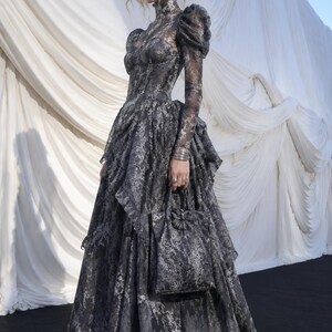 May include: A floor-length charcoal grey dress with a high neckline, long puff sleeves, and a corset-style bodice. The garment features an intricate, mottled pattern throughout and is paired with a matching handbag with a floral accent.