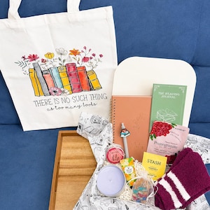 Może przedstawiać: Biała pł&oacute;cienna torba z kolorową ilustracją książek i kwiat&oacute;w oraz napisem "THERE IS NO SUCH THING as too many books." Na drewnianej tacy ułożono notatnik, dziennik czytelnika, herbatę, świecę oraz parę skarpetek.