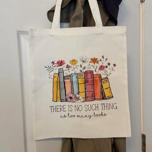 Może przedstawiać: Biała pł&oacute;cienna torba z kolorową ilustracją rzędu książek, z kt&oacute;rych wyrastają kwiaty. Tekst nadrukowany pod książkami brzmi: "THERE IS NO SUCH THING as too many books" i jest napisany mieszanką liter drukowanych oraz pisma odręcznego.