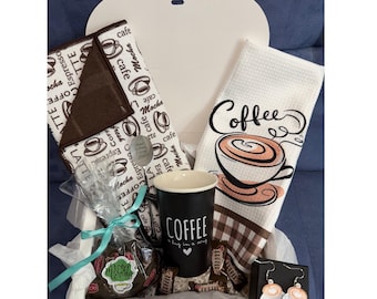 Może przedstawiać: Pudełko prezentowe zawierające czarny kubek z napisem COFFEE a hug in a mug, dwa białe ręczniki kuchenne w motywy kawowe, małą torebkę ze słodyczami przewiązaną jasnoniebieską wstążką oraz parę wiszących kolczyków w kształcie filiżanek kawy.