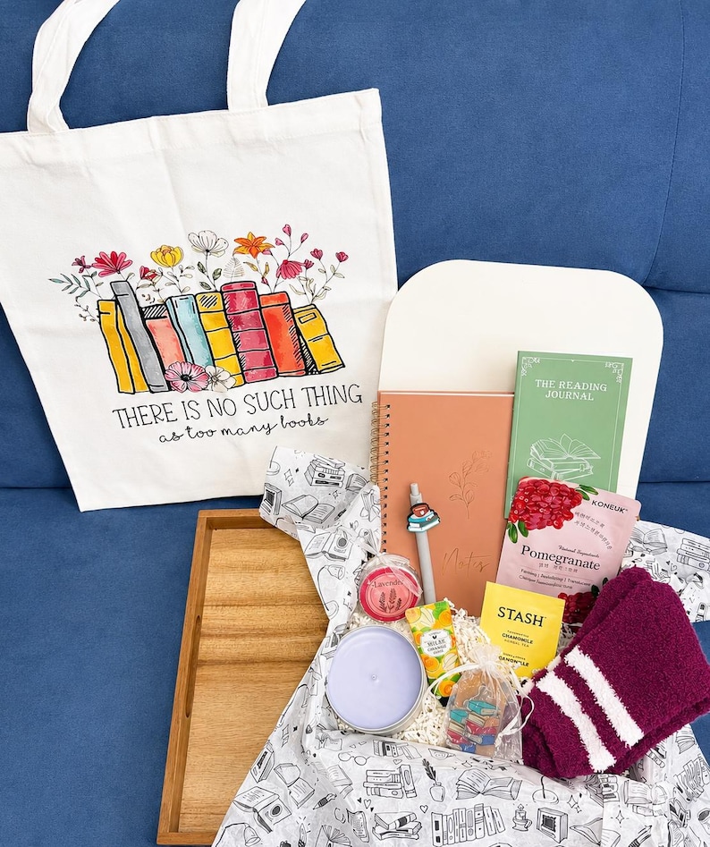 Może przedstawiać: Biała pł&oacute;cienna torba z kolorową ilustracją książek i kwiat&oacute;w oraz napisem "THERE IS NO SUCH THING as too many books." Na drewnianej tacy ułożono notatnik, dziennik czytelnika, herbatę, świecę oraz parę skarpetek.