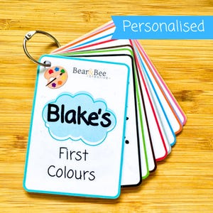 May include: A set of flashcards on a metal ring, featuring a white cover card with the text "Blake's First Colours" inside a light blue cloud graphic, a paint palette icon, and the brand name "Bear & Bee creative" in black text.