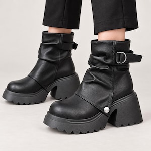 Könnte beinhalten: Schwarze Stiefeletten mit gerafftem Schaft, einer dicken Plateausohle mit gezacktem Profil und einem seitlichen Riemen mit silberfarbener Metallschnalle. Die Stiefel haben eine abgerundete Zehenpartie, einen silbernen Druckknopf und eine matte Oberfläche.