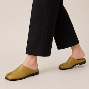 May include: A pair of olive green leather mules featuring a distinct split-toe design, a low black heel, and a smooth finish. The footwear is worn with black trousers, showcasing the slip-on style and the unique toe separation detail clearly.