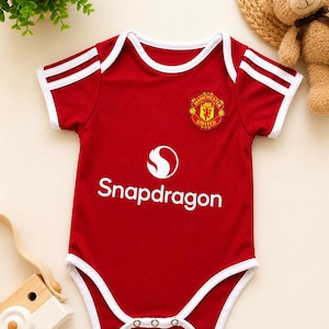 May include: A red baby bodysuit featuring white trim on the sleeves and leg openings, with two white stripes on each shoulder. The chest displays the Manchester United logo and the white text Snapdragon with its corresponding circular logo above it.