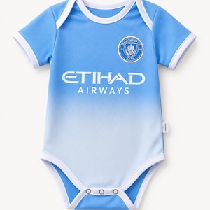 Op de afbeelding: Een lichtblauwe baby-romper met een wit kleurverloop aan de onderkant. Het kledingstuk toont het embleem van voetbalclub Manchester City en het Etihad Airways-logo in het wit. Het heeft korte mouwen, witte biezen langs de hals, mouwen en beenopeningen, en drie metalen drukknopen aan de onderkant.