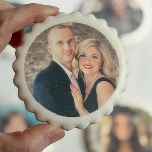 May include: A white, scalloped-edged cookie with a photo of a couple, held by a hand. The photo is surrounded by a white circle. The word "engagement" is written in a yellow rectangle above the cookie.