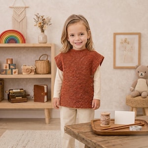 May include: A sleeveless, rust-colored knitted vest featuring a textured stitch pattern and two buttons on the side. The garment is worn over a long-sleeved, cream-colored shirt and light-colored trousers, creating a layered look.