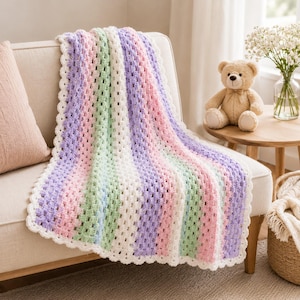 May include: A handmade crochet blanket featuring vertical stripes in pastel shades of pink, light purple, mint green, and white. The blanket has a textured granny stitch pattern and a scalloped white border edge, draped over a neutral sofa.