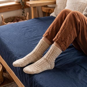 Peut inclure: Une paire de chaussettes beige mi-mollet avec un motif en tricot torsadé vertical et une texture légèrement mouchetée. Les chaussettes sont portées aux pieds, remontant au-dessus de la cheville, et présentent un bord côtelé pour un maintien ajusté.
