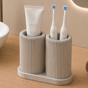Può includere: Un organizer da bagno grigio chiaro con due scomparti cilindrici a coste verticali su una base ovale. Uno scomparto contiene un tubetto di dentifricio Livora Dental Care, l'altro due spazzolini elettrici bianchi.