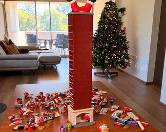 Könnte beinhalten: Ein hoher, rechteckiger Adventskalender in Form eines Schornsteins mit rotem Backsteinmuster und einer oben feststeckenden Weihnachtsmannfigur. Die Basis zeigt eine gewölbte Kaminöffnung. Ringsherum liegen verstreute Süßigkeiten und Schokolade.