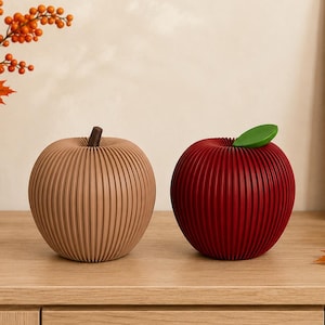 May include: Two decorative apple-shaped objects with a ribbed, accordion-style texture. One apple is light tan with a dark brown stem, and the other is deep red with a bright green leaf attached to the top. Both items have a rounded, spherical form.
