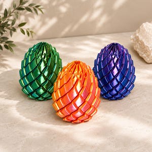 Pode incluir: Três objetos decorativos em forma de ovo com acabamento metálico e uma textura de escamas com padrão de losangos repetitivos. Um objeto é verde esmeralda, um é laranja brilhante e um é azul real profundo, todos com topo pontiagudo e base arredondada.