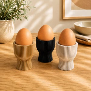 Può includere: Tre portauova nei colori beige, nero e bianco sporco, ciascuno caratterizzato da una texture a coste verticali e una base svasata. Ogni portauovo contiene un uovo marrone. I portauova sono disposti in fila su una superficie in legno.
