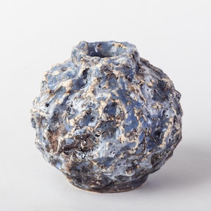 Könnte beinhalten: Eine kugelförmige Keramikvase mit einer stark texturierten, unebenen Oberfläche. Die Außenseite weist eine geschichtete Glasur in staubigen Blau-, Creme- und Dunkelbrauntönen auf, die ein raues, kraterartiges Aussehen mit einer kleinen Öffnung erzeugt.