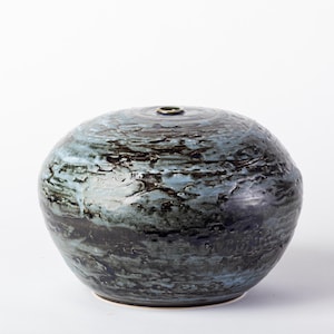 May include: A round ceramic vase with a low profile and a small circular opening at the top. The surface features a textured, mottled glaze in shades of deep black, dark blue, and hints of light blue, creating a layered, organic appearance.
