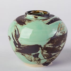 May include: A round ceramic vase with a glossy mint green glaze and bold, dark brown brushstroke patterns. The vessel features a short, narrow neck with a dark rim and a smooth, rounded body that tapers slightly toward a plain, unglazed base.