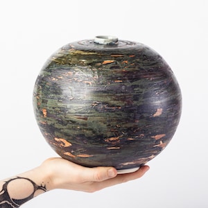 May include: A spherical ceramic vase with a dark, textured surface featuring mottled shades of forest green, deep purple, and metallic copper accents. The vase has a small, circular opening at the top with a light-colored, smooth rim.
