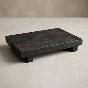 Può includere: Un rialzo rettangolare in legno con una finitura grigio carbone scuro e una trama visibile delle venature del legno. La piattaforma è sollevata da quattro piedini corti a forma di blocco su ogni angolo, offrendo una base stabile.