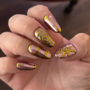 May include: Almond-shaped fingernails featuring a metallic pink base coat with raised gold chrome accents. The designs include a sun motif on the thumb, a spiral pattern on the middle finger, and abstract gold lines on the index, ring, and pinky nails.