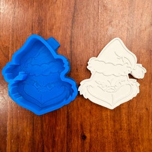 May include: A blue plastic cookie cutter and a white stamped dough cutout, both shaped like a character wearing a Santa hat with a frowning expression, furrowed eyebrows, and a wide, thin-lipped mouth, displaying the detailed facial features.
