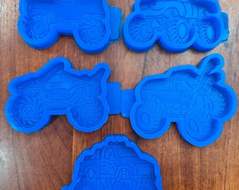 May include: A set of five blue plastic cookie cutters shaped like monster trucks. Each cutter features detailed impressions of large tires, truck bodies, and unique designs including a shark, a bull, and flames, designed for cutting dough shapes.
