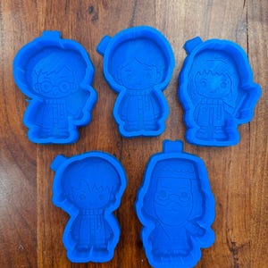 May include: Five bright blue silicone cookie cutters shaped like cartoon characters. The set includes figures with distinct hairstyles, scarves, and facial features, including one character with round glasses and another with a long beard and hat.