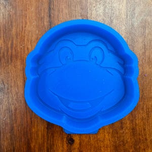 May include: A bright blue silicone mold featuring a raised, embossed design of a cartoon character face with large eyes, a wide nose, and a smiling mouth. The mold has a rounded, circular shape with a small tab at the bottom edge for easy handling.