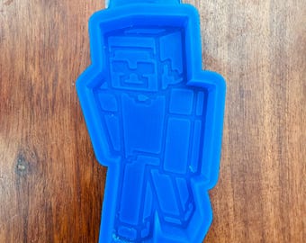 May include: A bright blue silicone mold featuring the recessed, blocky, pixelated silhouette of a character with a square head, rectangular torso, and limbs, designed for casting shapes in the likeness of a video game figure.