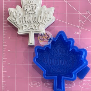 Könnte beinhalten: Ein weißer Abguss in Ahornblattform und eine blaue Silikonform, beide mit dem geprägten Text "HAPPY CANADA DAY!". Die Gegenstände liegen auf einer rosa Schneidematte mit sichtbaren Rasterlinien und Maßen in Zoll.