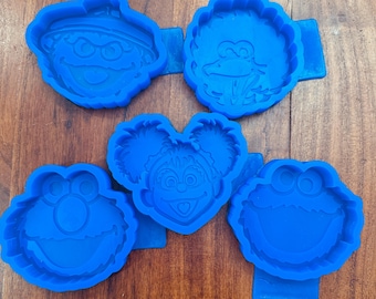 May include: A set of five blue silicone molds featuring embossed character faces from Sesame Street, including Elmo, Cookie Monster, Big Bird, Abby Cadabby, and Oscar the Grouch, arranged in a cluster on a flat surface.