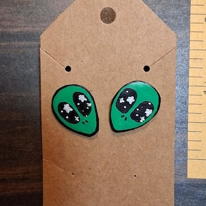 May include: A pair of green alien head stud earrings with black eyes featuring white star patterns, displayed on a brown cardboard backing card next to a ruler showing measurements in inches for scale and size reference.