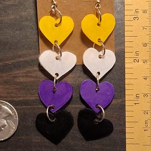 May include: A pair of dangle earrings featuring four stacked heart-shaped charms in yellow, white, purple, and black, suspended from gold-tone hooks. A ruler shows the earrings are approximately 3.5 inches long, next to a US quarter coin.