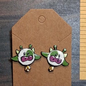 May include: A pair of stud earrings featuring a cartoon cow design with white and green patches, a purple snout, and a small gold-colored bell charm hanging from the bottom, displayed on a brown cardboard jewelry backing card.