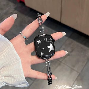 Peut inclure: Une montre connectée noire avec un écran numérique affichant l'heure 13:14 et des graphismes d'étoiles argentées. La montre est dotée d'un bracelet à maillons en métal argenté orné de petites breloques en forme d'étoile et de croissant de lune.