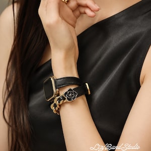 May include: A black leather watch band for a smartwatch featuring a gold-tone metal chain link section and a black flower charm. The band wraps around the wrist in a double-loop style with gold-tone hardware and a secure buckle closure.